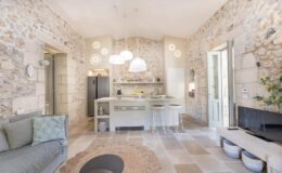 Modern open-concept kitchen and living area featuring natural stone walls, contemporary furniture, and stylish pendant lighting. The kitchen includes a central island with seating, sleek appliances, and decorative accents, while a cozy seating area showcases a patterned cushion and a round rug.