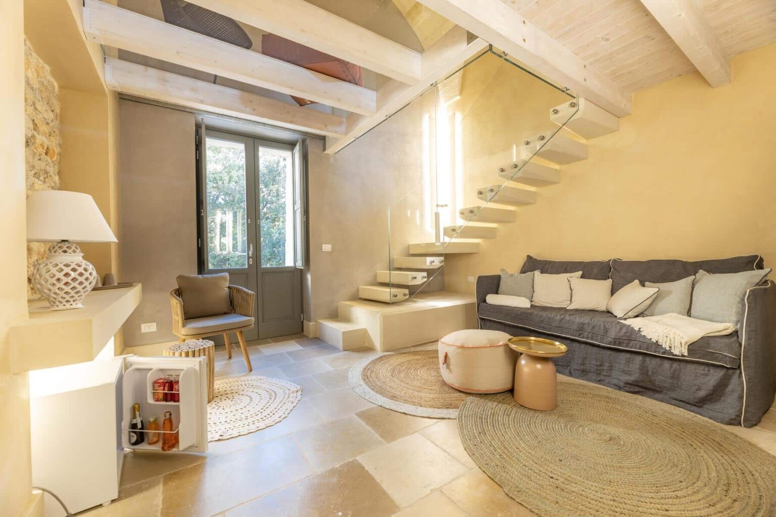 Cozy living room featuring a gray sofa, circular rugs, and a stylish staircase, illuminated by natural light from large windows. The space has a modern yet inviting design with neutral tones and decorative elements. | Only on SopranoVillas