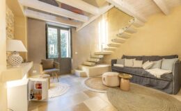 Cozy living room featuring a gray sofa, circular rugs, and a stylish staircase, illuminated by natural light from large windows. The space has a modern yet inviting design with neutral tones and decorative elements.