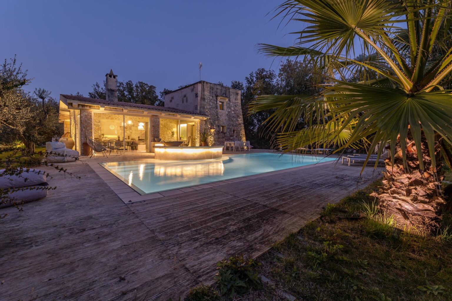 Beautiful outdoor pool area at dusk, featuring a modern swimming pool surrounded by lush greenery and illuminated by soft lighting, with a rustic stone building in the background. Ideal for relaxation and entertaining guests. | Only on SopranoVillas