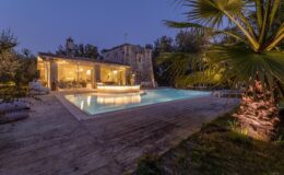 Beautiful outdoor pool area at dusk, featuring a modern swimming pool surrounded by lush greenery and illuminated by soft lighting, with a rustic stone building in the background. Ideal for relaxation and entertaining guests.