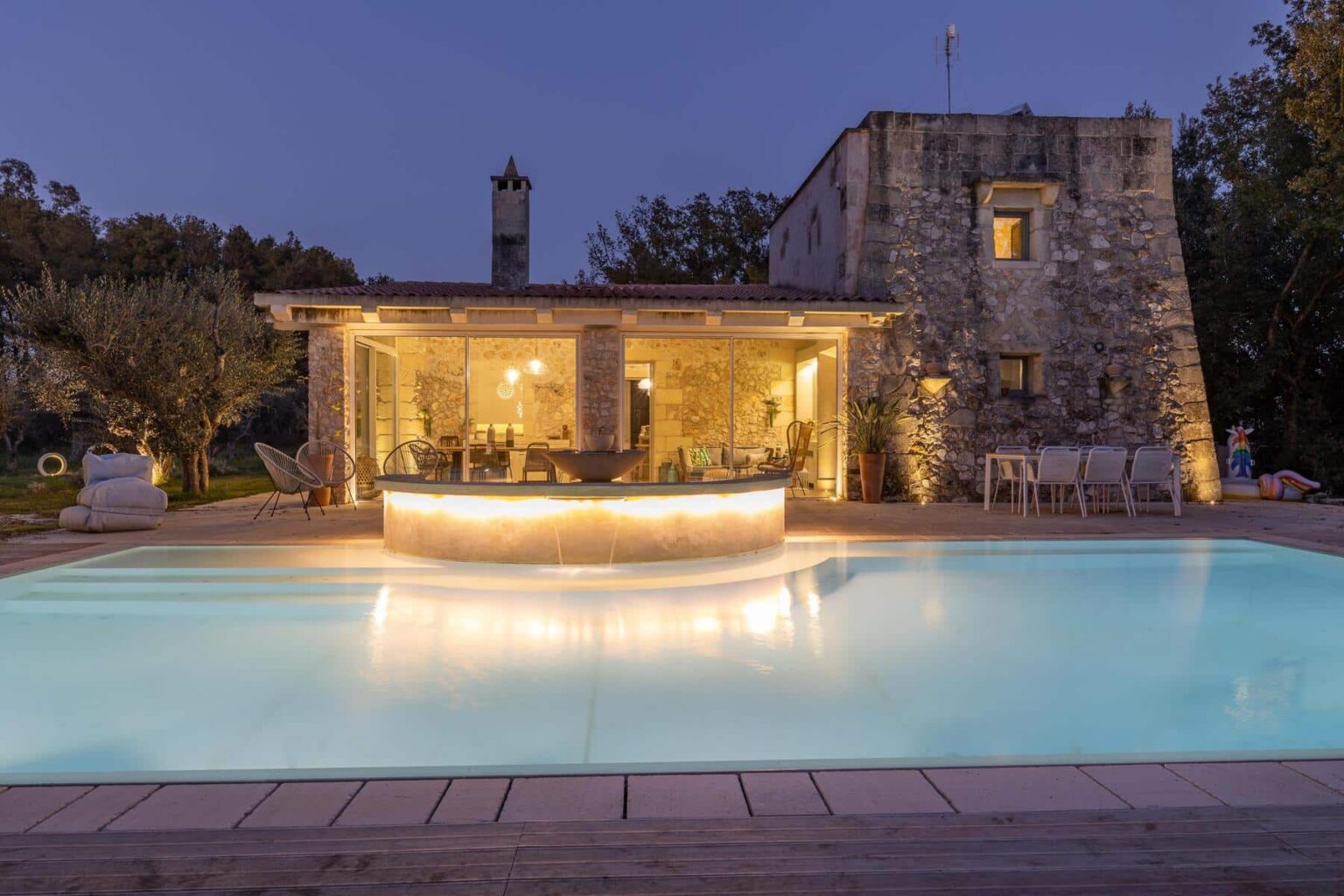 Modern stone villa with a glowing pool and outdoor seating area, surrounded by olive trees, showcasing a cozy interior with large glass windows at dusk. | Only on SopranoVillas
