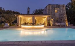 Modern stone villa with a glowing pool and outdoor seating area, surrounded by olive trees, showcasing a cozy interior with large glass windows at dusk.