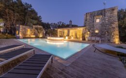 Luxury villa with a stunning pool area illuminated at dusk, surrounded by stone walls and lush trees, perfect for relaxation and outdoor gatherings.