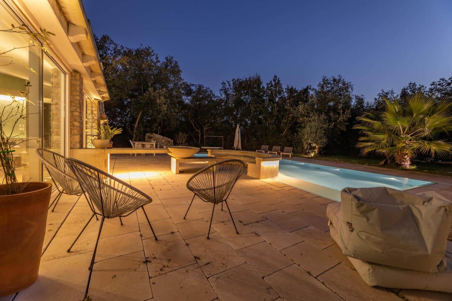 Cozy outdoor seating area with modern chairs beside a well-lit swimming pool at dusk, surrounded by lush greenery and a serene atmosphere. | Only on SopranoVillas