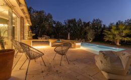 Cozy outdoor seating area with modern chairs beside a well-lit swimming pool at dusk, surrounded by lush greenery and a serene atmosphere.