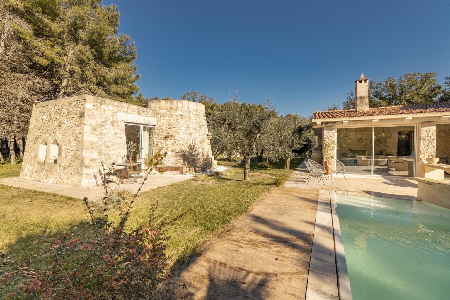 A picturesque stone house with a modern design, surrounded by olive trees and a swimming pool, set against a clear blue sky. The outdoor area features a wooden pathway and a cozy seating arrangement, ideal for relaxation and enjoying nature. | Only on SopranoVillas