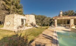 A picturesque stone house with a modern design, surrounded by olive trees and a swimming pool, set against a clear blue sky. The outdoor area features a wooden pathway and a cozy seating arrangement, ideal for relaxation and enjoying nature.