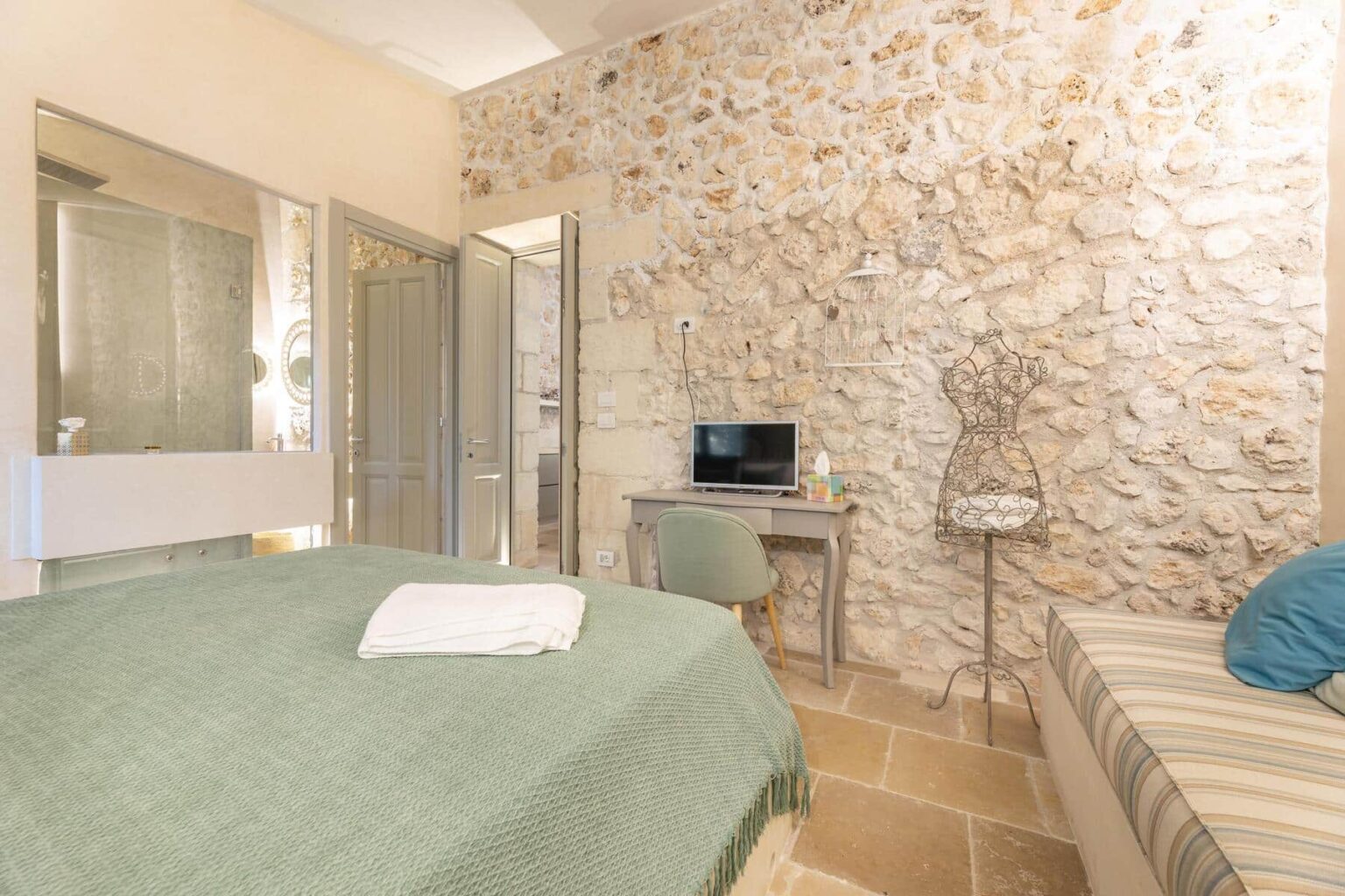 Cozy bedroom featuring a stone accent wall, a comfortable green bedspread, a small desk with a TV, and a stylish mannequin display. Ideal for relaxation and modern decor. | Only on SopranoVillas