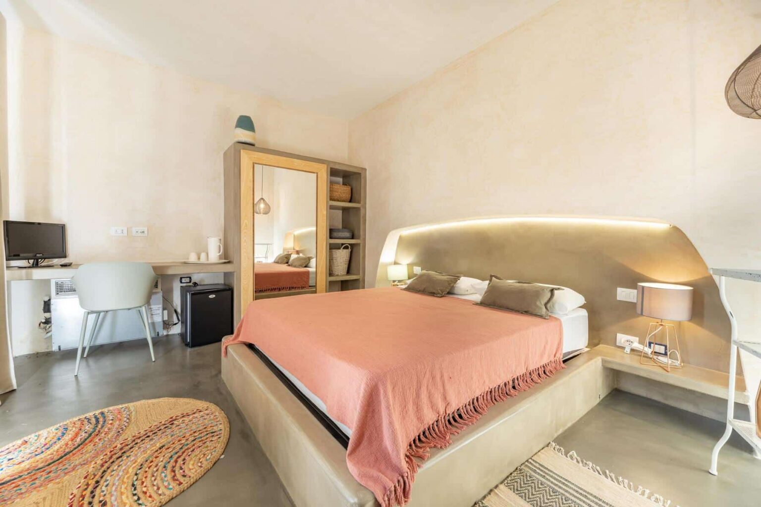 Cozy and modern bedroom featuring a queen-sized bed with a coral blanket, minimalist decor, a workspace with a desk and chair, a small refrigerator, and a mirror. Soft lighting enhances the warm ambiance, complemented by a decorative rug on the polished concrete floor. | Only on SopranoVillas