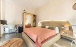 Cozy and modern bedroom featuring a queen-sized bed with a coral blanket, minimalist decor, a workspace with a desk and chair, a small refrigerator, and a mirror. Soft lighting enhances the warm ambiance, complemented by a decorative rug on the polished concrete floor.