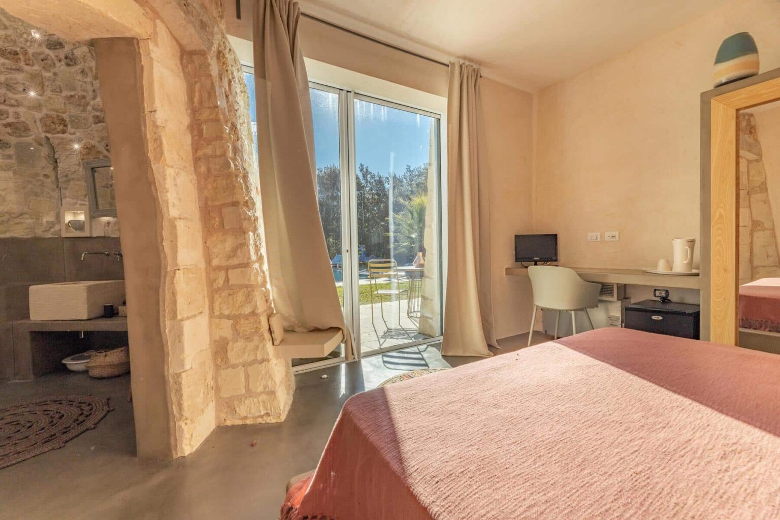 Cozy bedroom with a pink bedspread, featuring a stone wall and large glass doors leading to a patio. The room includes a desk with a chair and a small computer, along with a stylish decorative mirror. Sunlight streams in, highlighting the modern and rustic decor. | Only on SopranoVillas