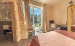Cozy bedroom with a pink bedspread, featuring a stone wall and large glass doors leading to a patio. The room includes a desk with a chair and a small computer, along with a stylish decorative mirror. Sunlight streams in, highlighting the modern and rustic decor.