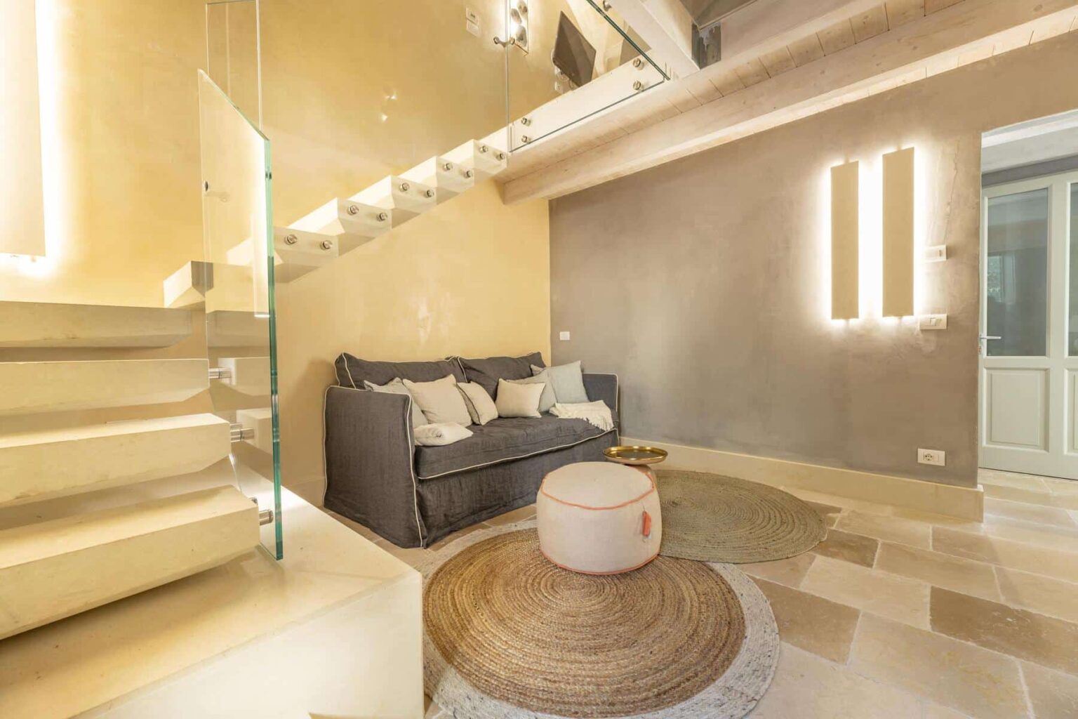 Modern living room interior featuring a gray sofa, round rug, and stylish lighting, with a staircase in the background. The space is designed with neutral tones and contemporary decor elements, creating a cozy and inviting atmosphere. | Only on SopranoVillas