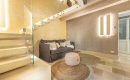 Modern living room interior featuring a gray sofa, round rug, and stylish lighting, with a staircase in the background. The space is designed with neutral tones and contemporary decor elements, creating a cozy and inviting atmosphere.