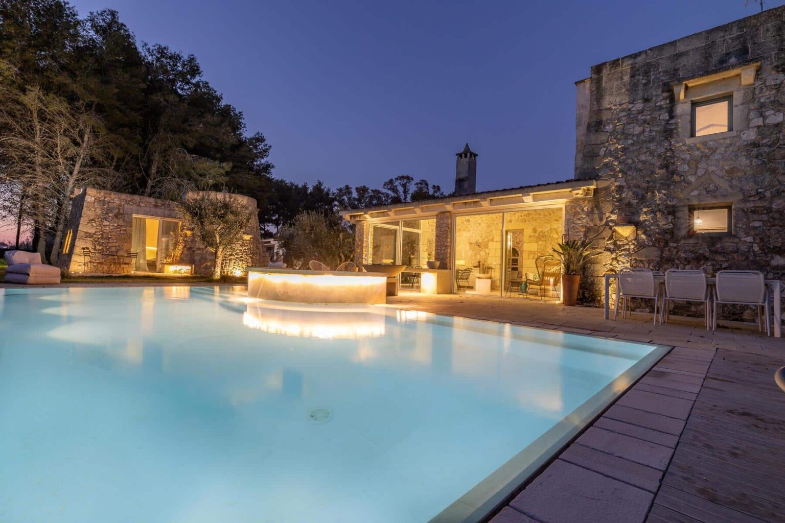 Luxury villa with a glowing pool and stone architecture, surrounded by trees at twilight, featuring a cozy outdoor seating area and modern furnishings. | Only on SopranoVillas