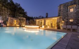 Luxury villa with a glowing pool and stone architecture, surrounded by trees at twilight, featuring a cozy outdoor seating area and modern furnishings.