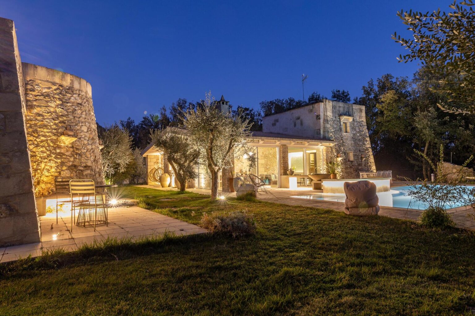 Charming villa at dusk with illuminated garden, stone architecture, and a serene swimming pool surrounded by olive trees. Ideal for a relaxing getaway. | Only on SopranoVillas