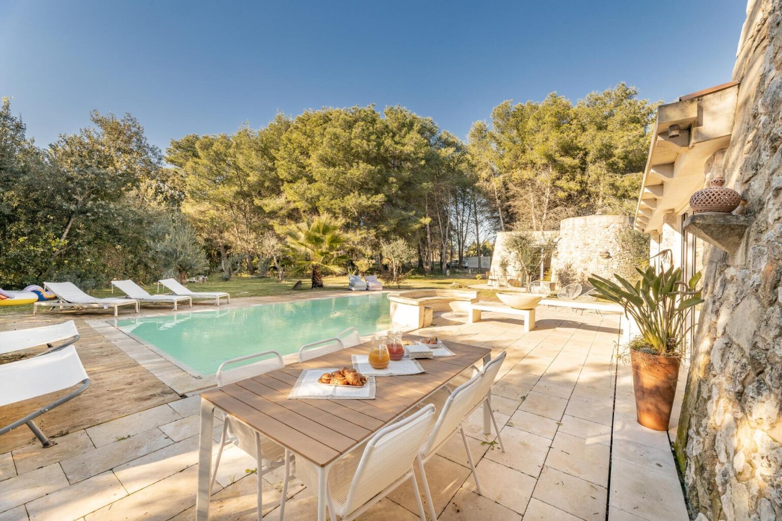 Luxurious outdoor pool area featuring a wooden deck, sun loungers, and a dining table set with pastries and drinks, surrounded by lush greenery and a serene landscape. Ideal for relaxation and entertaining. | Only on SopranoVillas