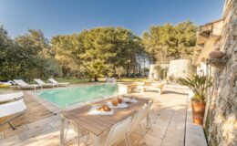 Luxurious outdoor pool area featuring a wooden deck, sun loungers, and a dining table set with pastries and drinks, surrounded by lush greenery and a serene landscape. Ideal for relaxation and entertaining.