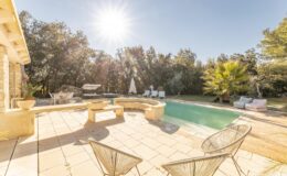 Luxury villa with a sunlit swimming pool, surrounded by lush greenery and outdoor seating, perfect for relaxation and leisure.
