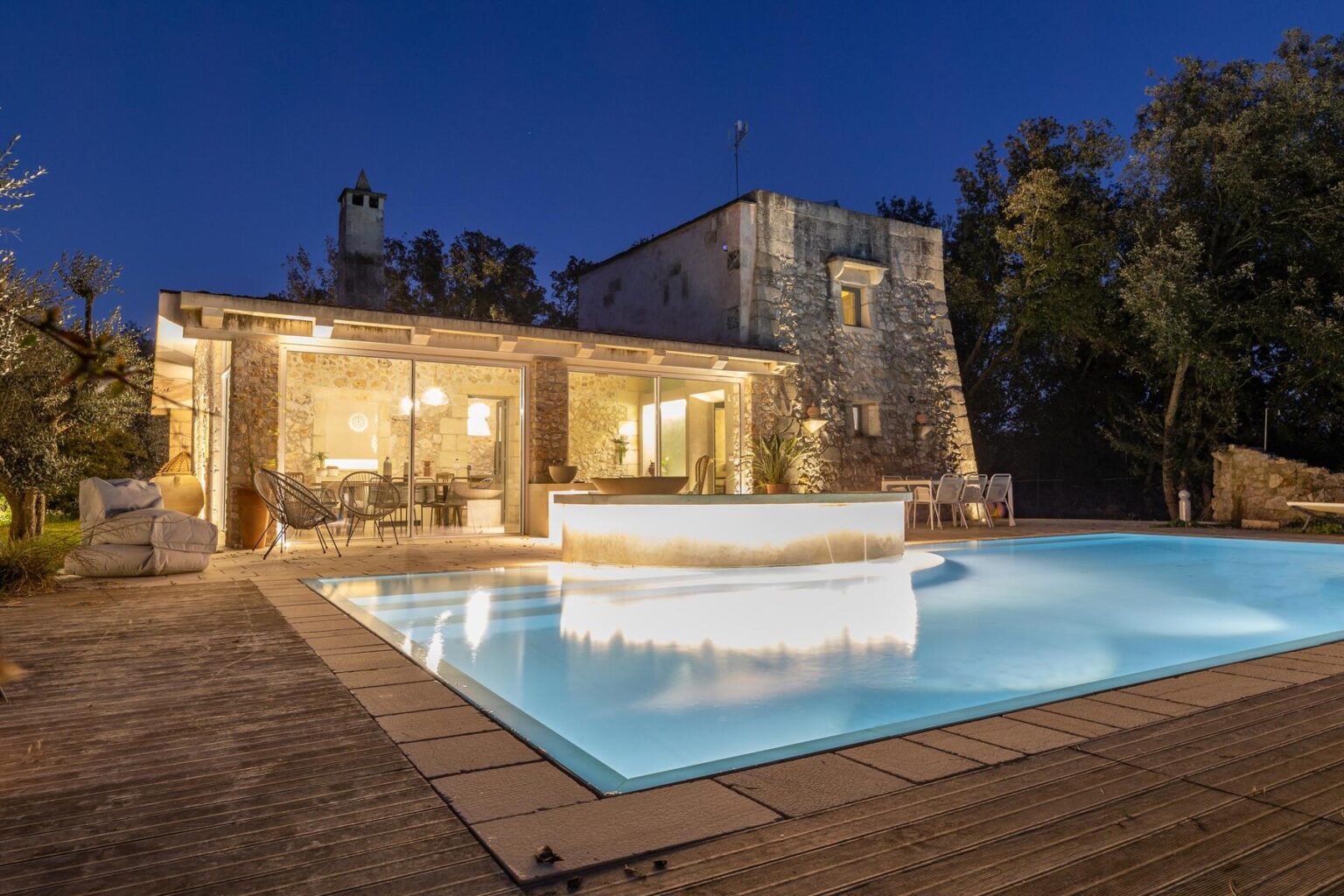 Luxury villa with a glowing swimming pool at night, surrounded by lush greenery and stone architecture, showcasing a cozy outdoor seating area. Ideal for relaxation and entertainment. | Only on SopranoVillas