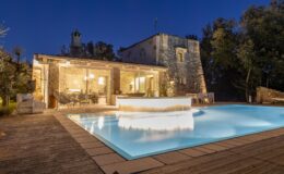 Luxury villa with a glowing swimming pool at night, surrounded by lush greenery and stone architecture, showcasing a cozy outdoor seating area. Ideal for relaxation and entertainment.