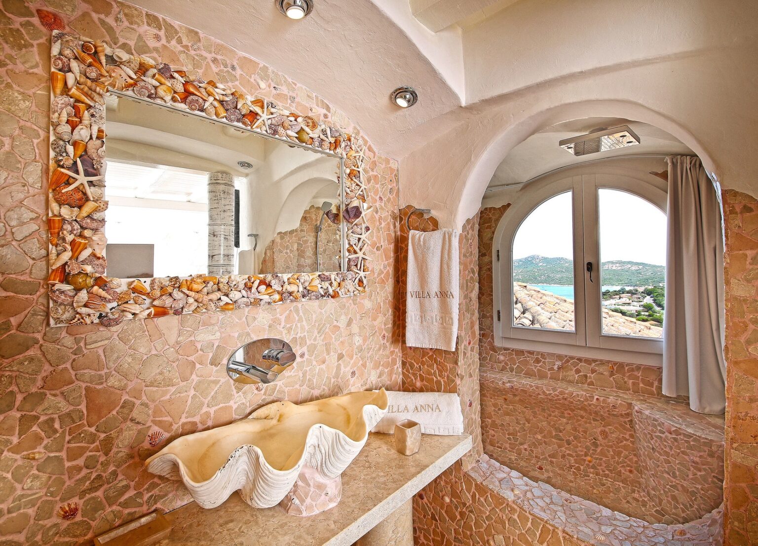 Elegant bathroom featuring a unique shell-decorated mirror, a natural stone sink, and a picturesque view through the arched window. The walls are adorned with pink stone tiles, creating a serene atmosphere. Ideal for luxury villa design inspiration. | Only on SopranoVillas