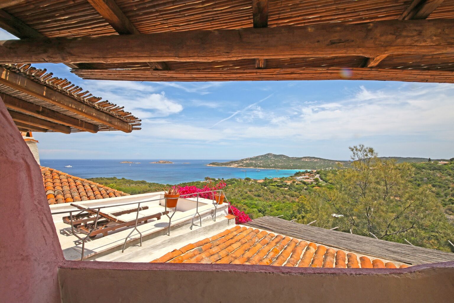 Scenic view from a terrace overlooking a coastal landscape with turquoise waters, lush greenery, and distant islands under a blue sky, featuring terracotta roofing and potted flowers. | Only on SopranoVillas