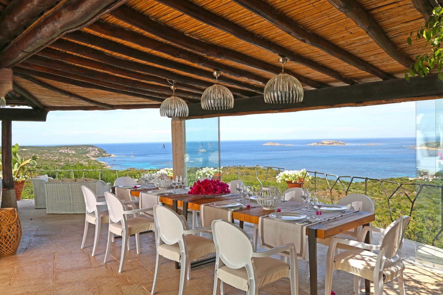 Scenic outdoor dining area with panoramic ocean views, featuring a wooden table set for a meal, elegant glassware, and stylish white chairs under a thatched roof with decorative pendant lights. Lush greenery and a coastal landscape enhance the tranquil atmosphere. | Only on SopranoVillas