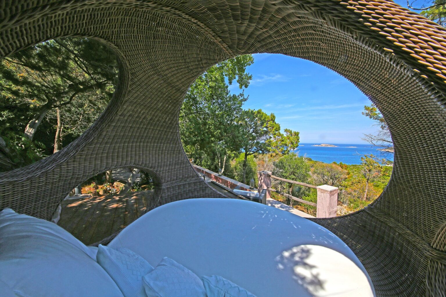 Cozy outdoor seating area with a round wicker design, offering a stunning view of the ocean and lush greenery under a clear blue sky. Perfect for relaxation and enjoying nature. | Only on SopranoVillas
