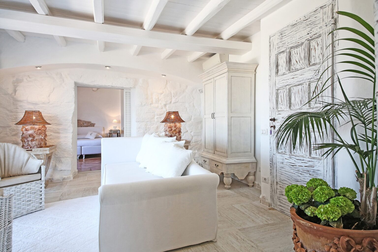 Bright and airy living room featuring a white sofa, decorative lamps, and a lush green plant, with rustic stone walls and wooden beams, creating a cozy and inviting atmosphere. | Only on SopranoVillas
