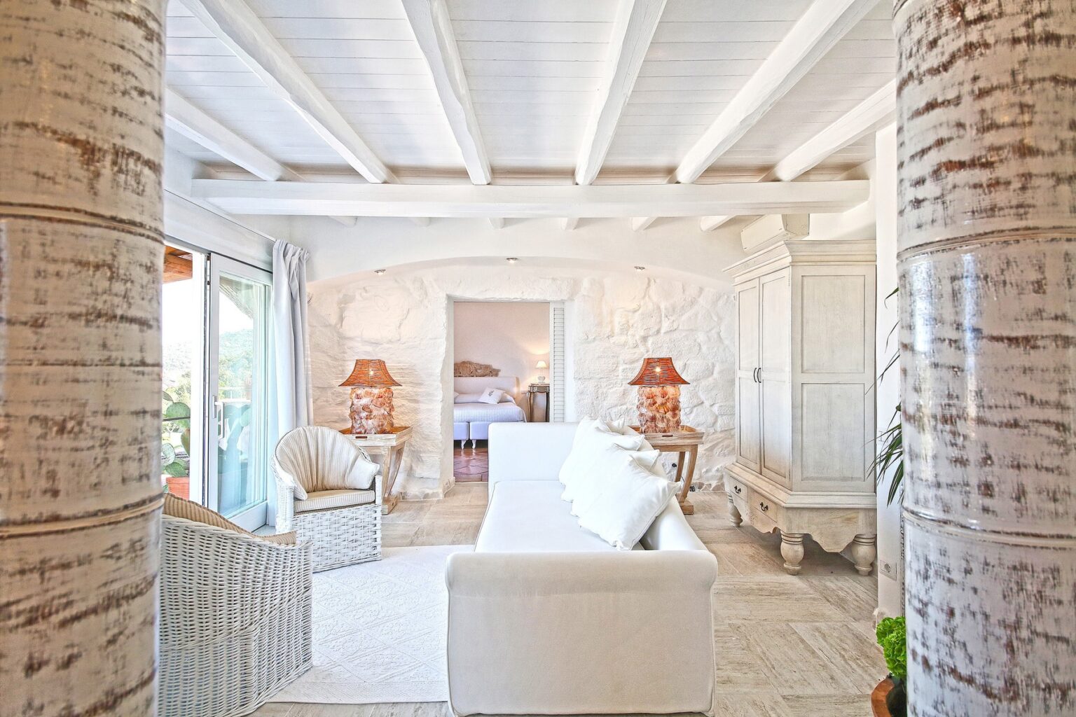 Bright and airy living room featuring white furniture, textured columns, and elegant decor. The space is enhanced by natural light, showcasing a cozy atmosphere with stylish lamps and a view of a serene outdoor area. Ideal for a relaxing retreat. | Only on SopranoVillas
