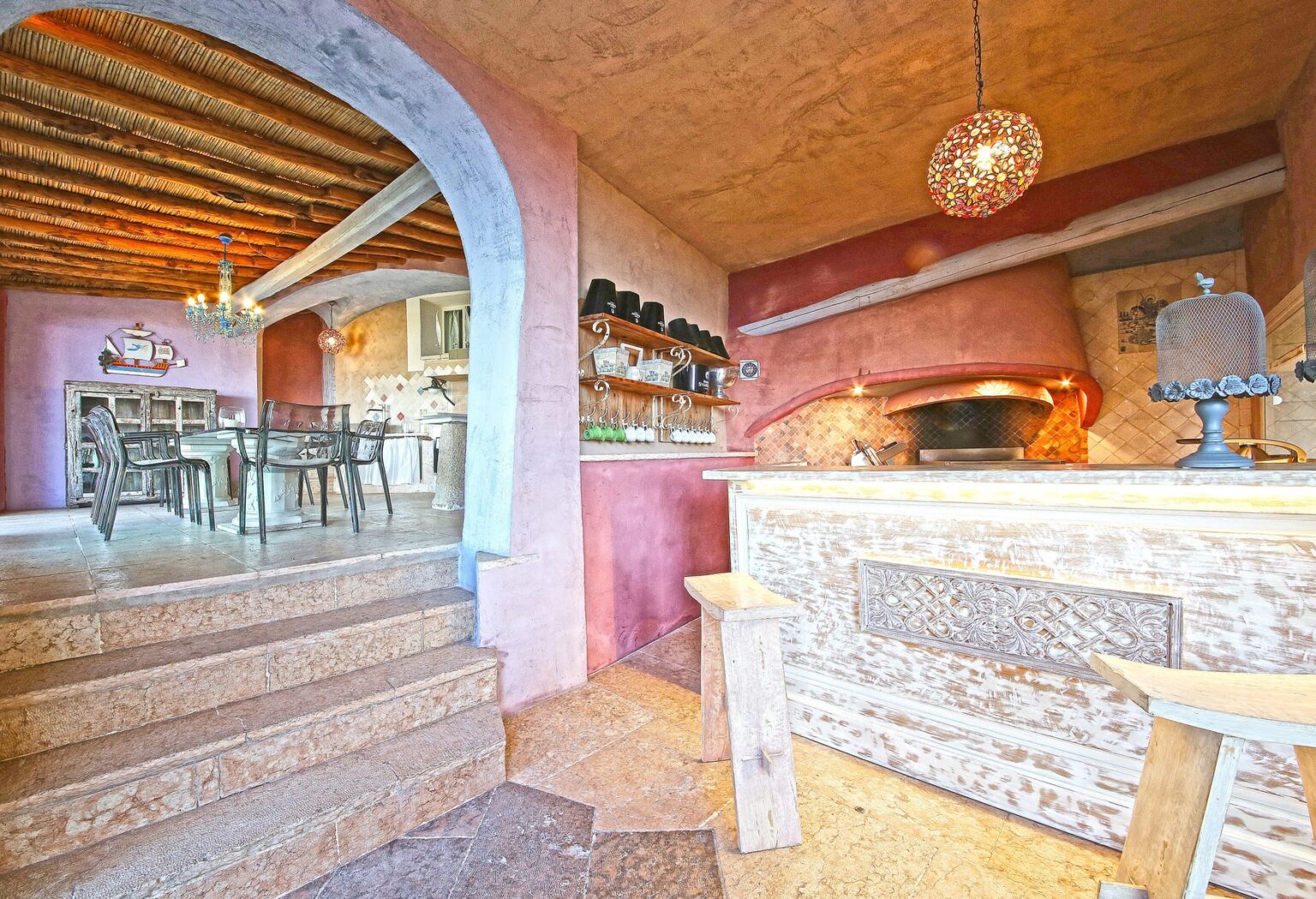 Cozy rustic kitchen interior featuring a stone pizza oven, wooden accents, and a dining area with a large table. The walls are adorned with warm colors, and a chandelier adds a charming touch to the space. | Only on SopranoVillas