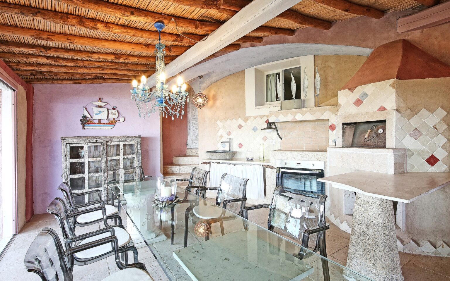 Stylish dining area featuring rustic decor, a wooden beam ceiling, a glass table, and comfortable seating, complemented by a modern kitchen and a charming fireplace. Perfect for cozy gatherings and entertaining guests. | Only on SopranoVillas