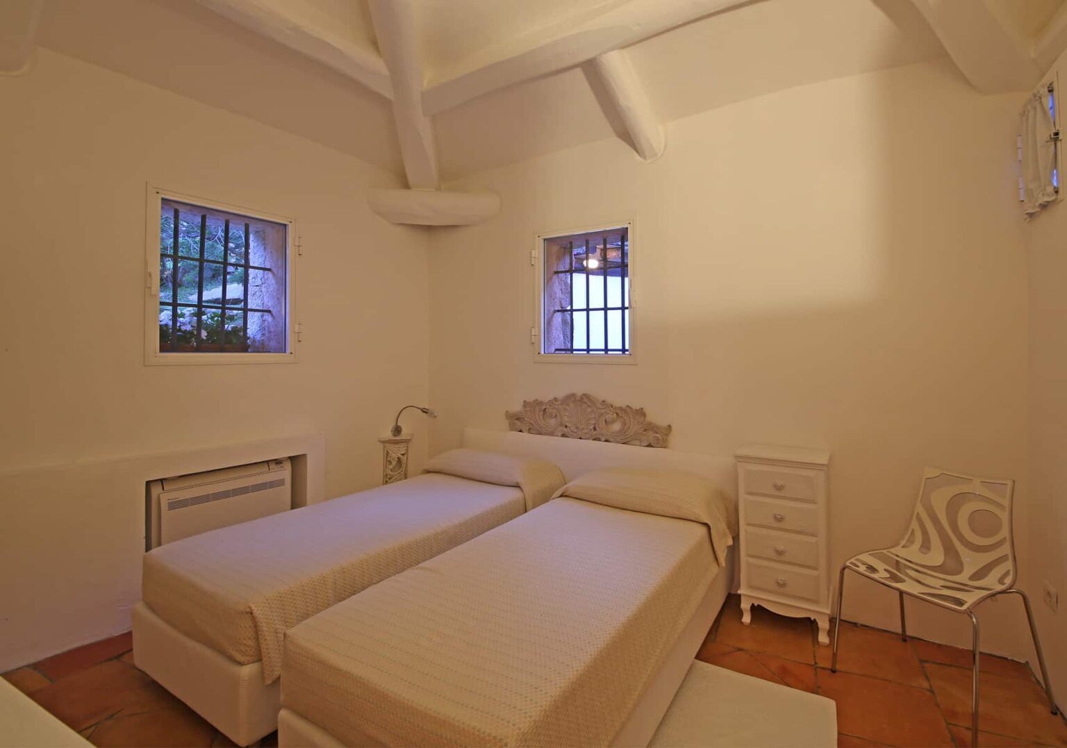 Cozy bedroom featuring two single beds, white walls, and natural light from windows. Includes a bedside table and a decorative chair, creating a minimalist and inviting atmosphere. Perfect for relaxation and comfort. | Only on SopranoVillas