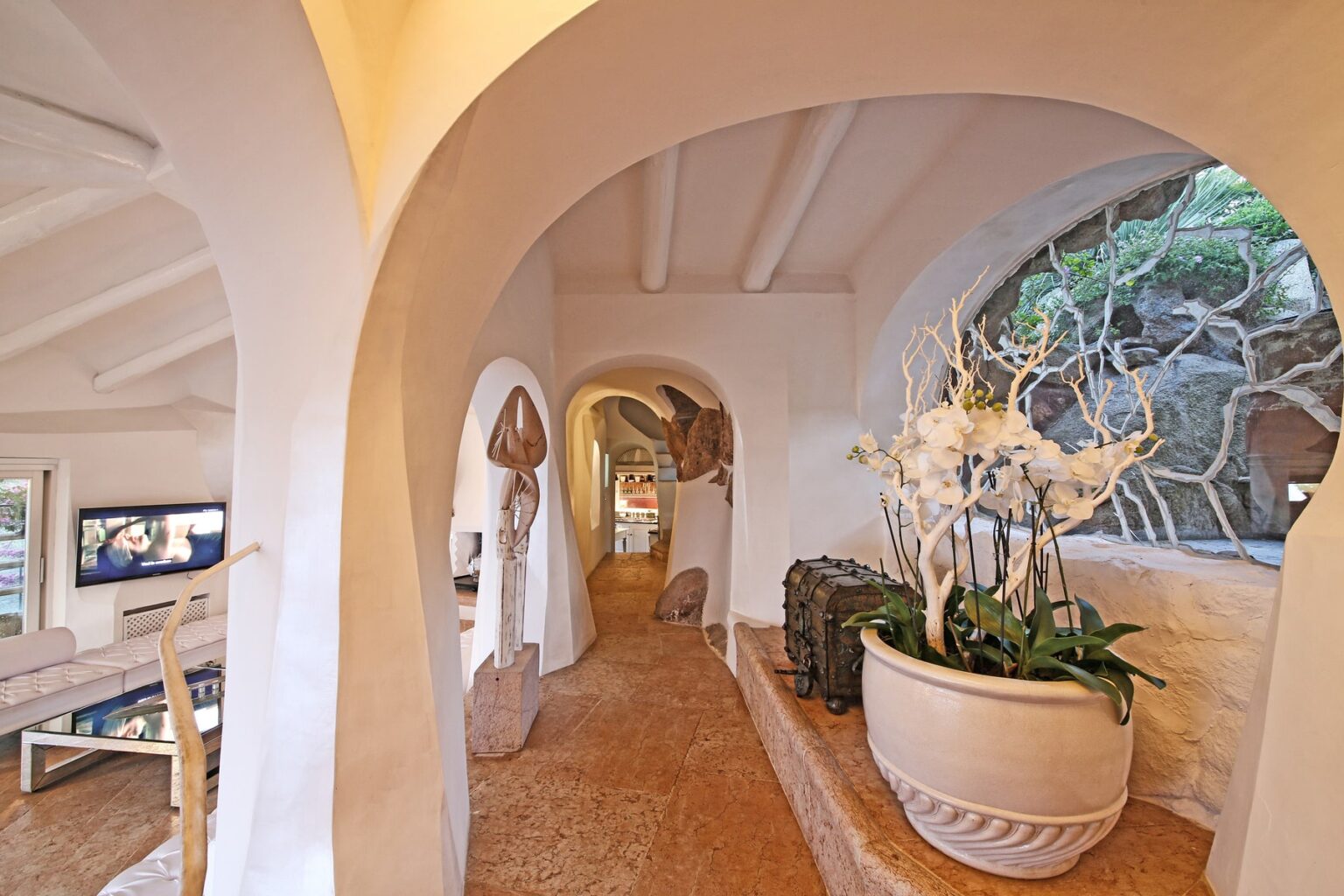 Interior view of a modern home showcasing an arched hallway with white walls, natural light, and decorative potted plants. The design features a blend of organic shapes and earthy tones, creating a serene atmosphere. | Only on SopranoVillas