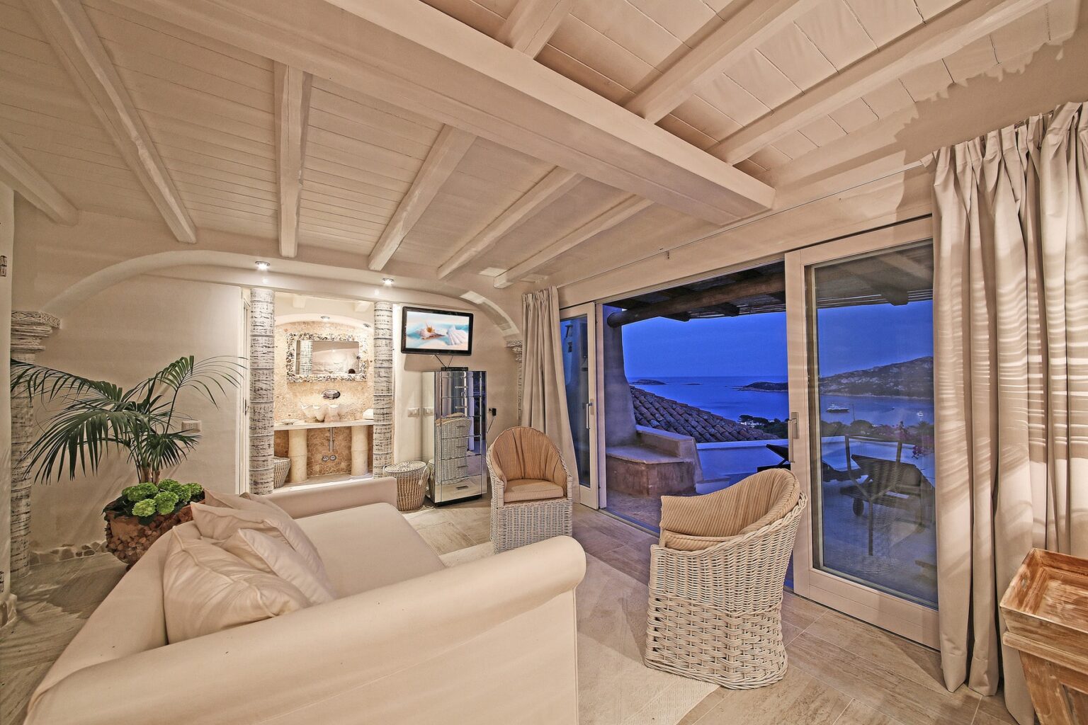 Cozy living room with white wooden beams, a comfortable sofa, and ocean views through large glass doors, featuring modern decor and warm lighting. | Only on SopranoVillas