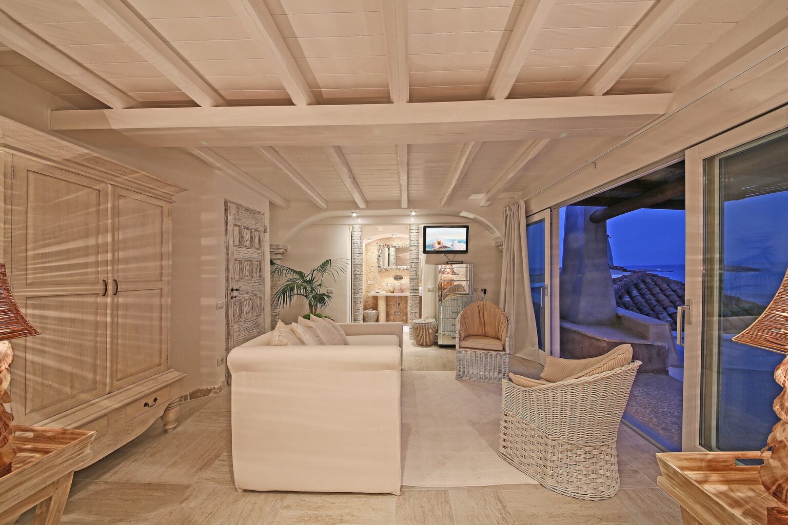 Cozy beach house interior featuring whitewashed walls, a comfortable sofa, wicker chairs, and large windows offering a view of the ocean at dusk. | Only on SopranoVillas