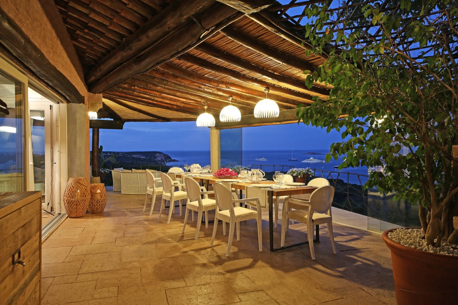 Elegant outdoor dining area with a large table and white chairs, surrounded by lush greenery, featuring pendant lighting and a stunning ocean view at dusk. Perfect for entertaining and enjoying the sunset. | Only on SopranoVillas
