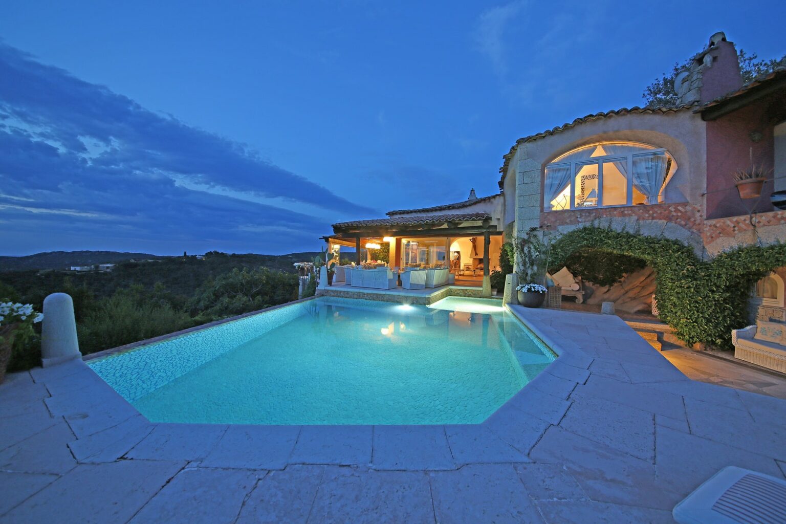 Luxury villa with a stunning pool at dusk, surrounded by lush greenery and scenic views. The warm glow from the interior lights creates an inviting atmosphere, perfect for relaxation and outdoor entertaining. | Only on SopranoVillas