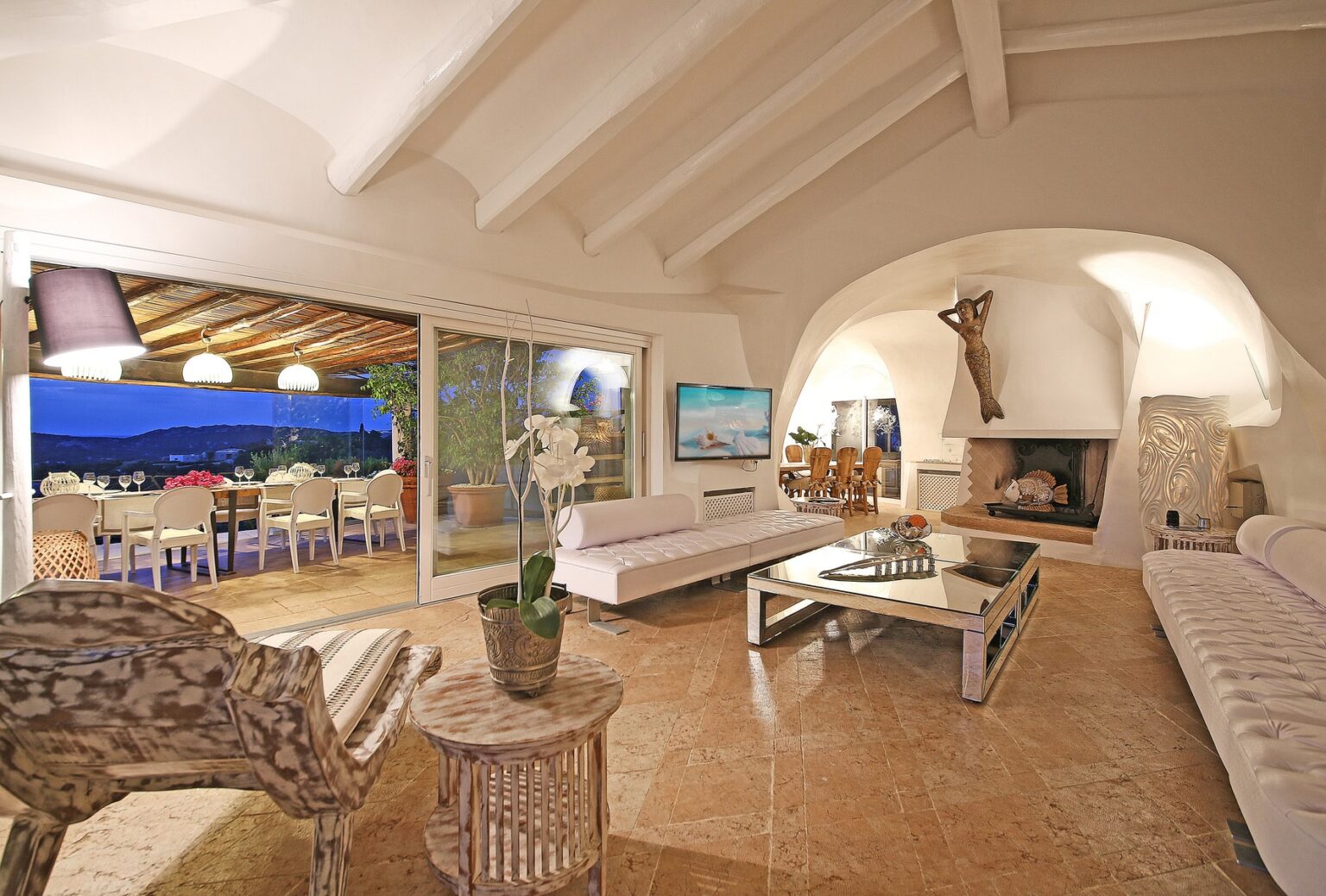 Modern living room with high ceilings, featuring elegant white furniture, a cozy fireplace, and a large window overlooking an outdoor dining area with ocean views. Decorative elements include a stylish lamp and a potted orchid. | Only on SopranoVillas