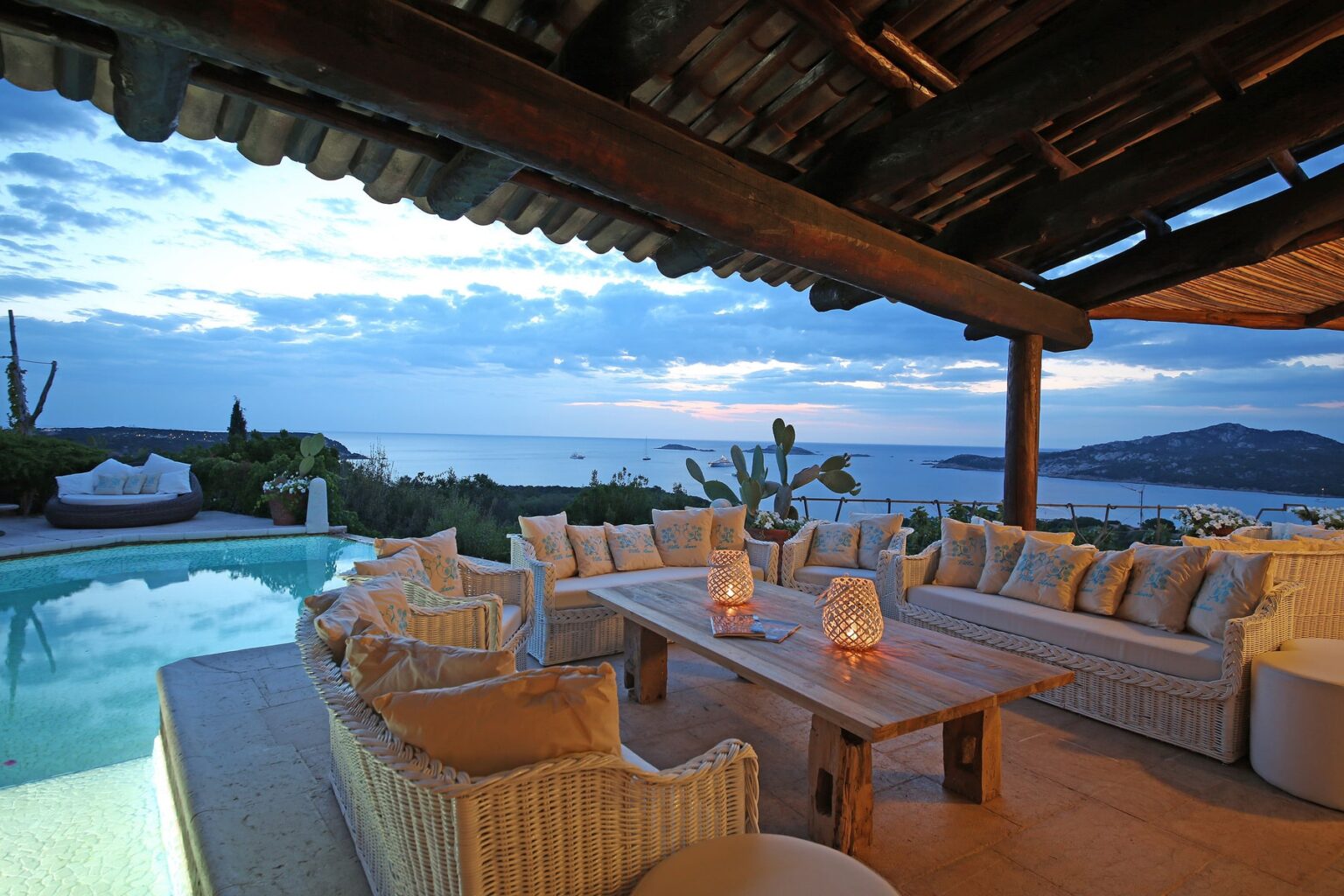 Luxurious outdoor seating area overlooking a serene ocean view at sunset, featuring a wooden table surrounded by white wicker sofas adorned with decorative pillows, and illuminated by stylish lanterns beside a tranquil swimming pool. | Only on SopranoVillas