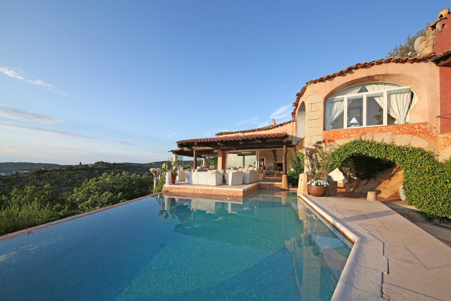 Luxurious villa with a stunning infinity pool overlooking lush hills, featuring a spacious outdoor seating area and elegant architecture under a clear blue sky. Perfect for relaxation and scenic views. | Only on SopranoVillas