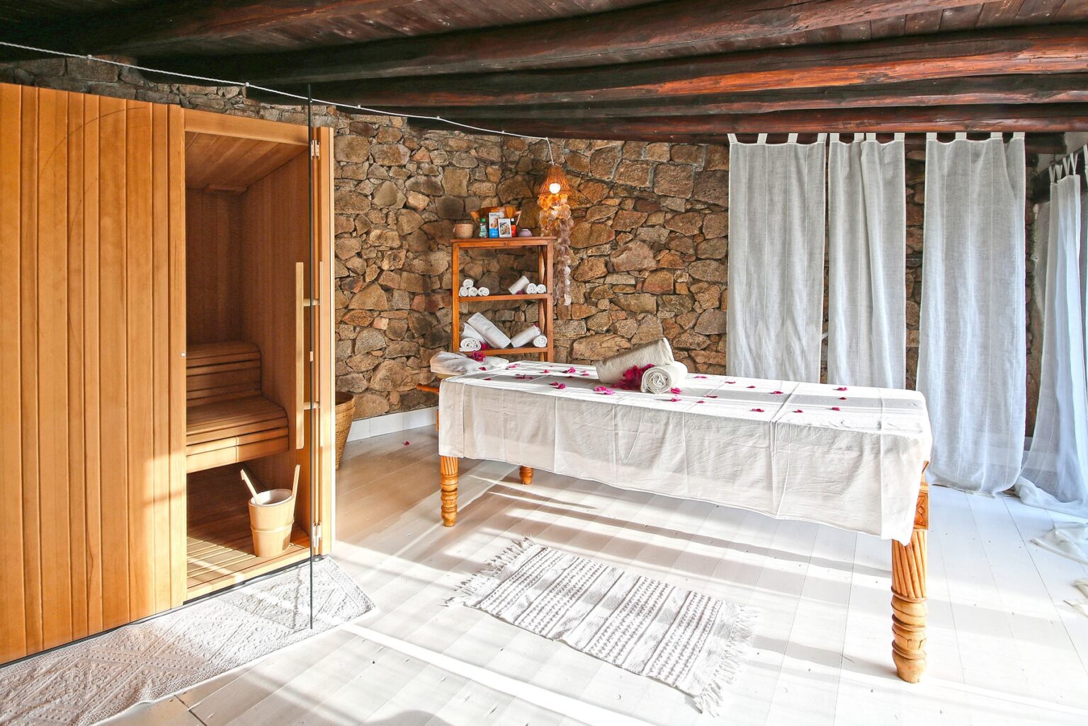 Relaxing spa room featuring a wooden sauna, a massage table adorned with rose petals, and neatly arranged towels on a rustic shelf. The space is illuminated by soft natural light filtering through sheer curtains, creating a serene atmosphere for wellness and relaxation. | Only on SopranoVillas