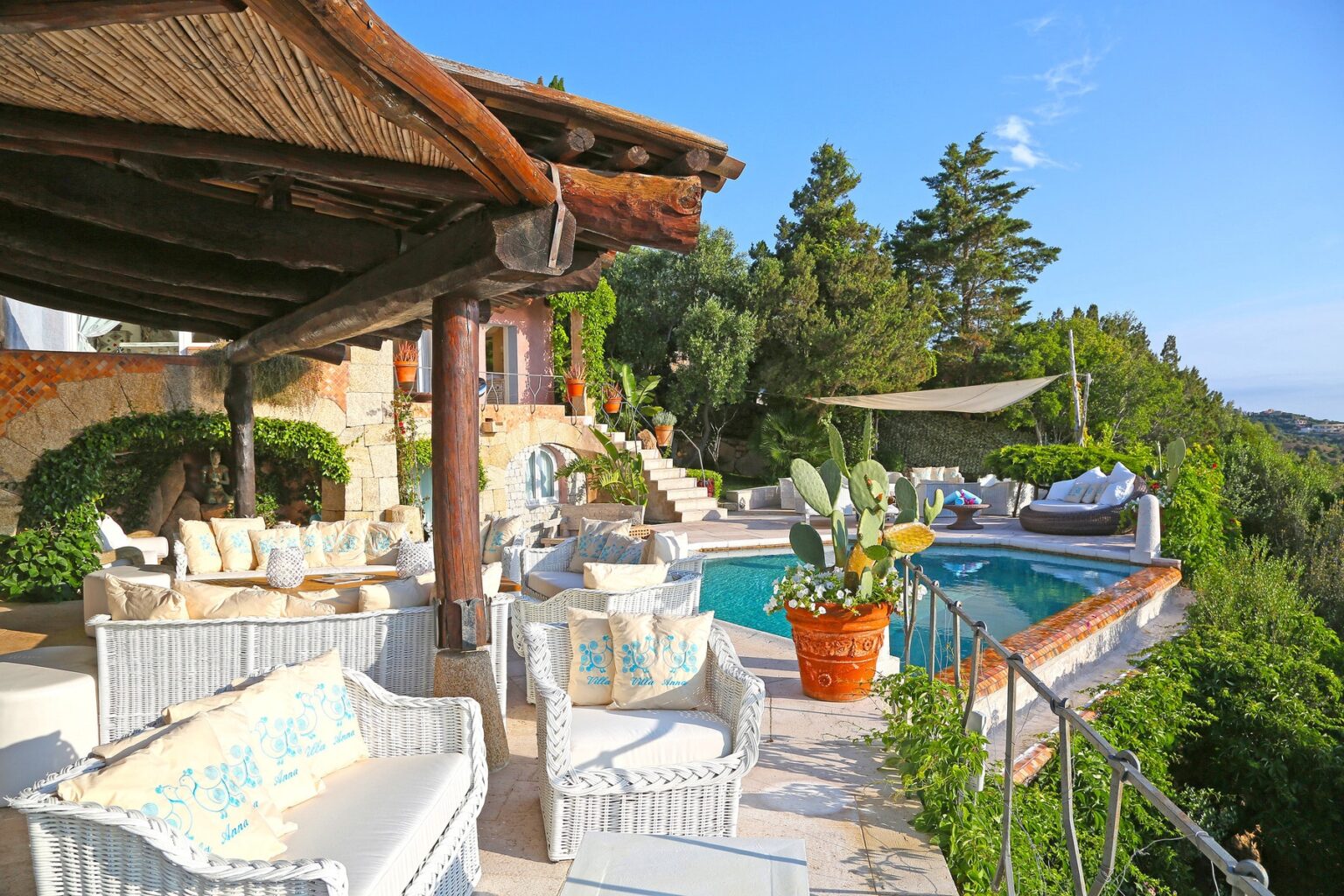 Luxurious outdoor patio with white wicker furniture and a swimming pool, surrounded by lush greenery and trees, ideal for relaxation and entertaining in a scenic setting. | Only on SopranoVillas