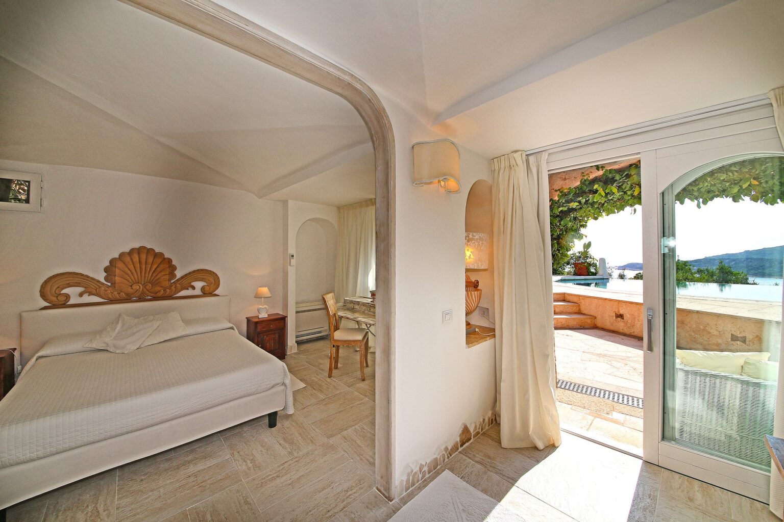 Cozy bedroom with a stylish shell-shaped headboard, featuring a comfortable bed, wooden nightstand, and a small study area. The room opens to a scenic outdoor space with large glass doors, offering views of a tranquil landscape and a swimming pool surrounded by greenery. | Only on SopranoVillas