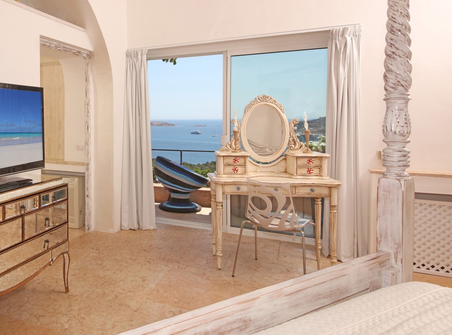 Elegant bedroom interior featuring a vintage vanity with a mirror, a TV, and large windows showcasing a stunning ocean view. Soft lighting and neutral tones create a serene atmosphere. | Only on SopranoVillas