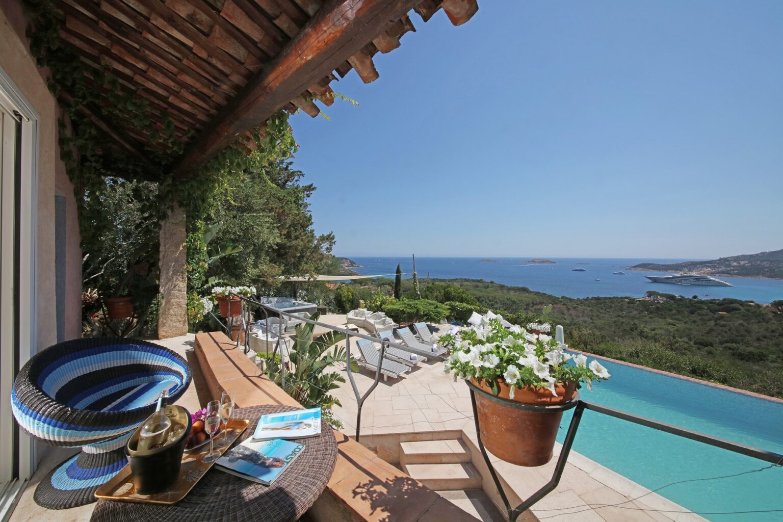 Scenic view from a villa terrace overlooking a tranquil sea and lush greenery, featuring a wooden table with decorative plants and a swimming pool in the foreground. | Only on SopranoVillas