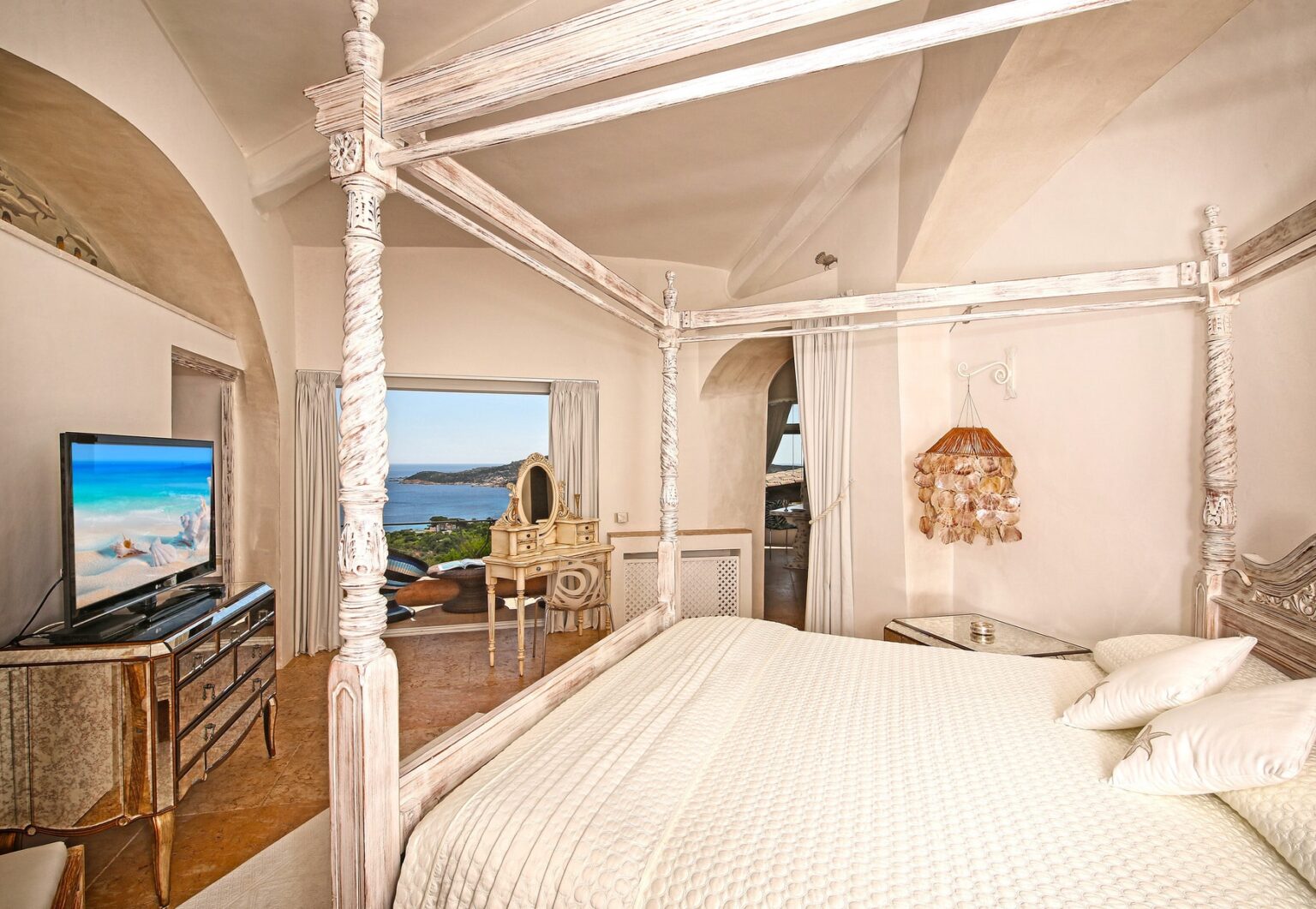 Elegant bedroom featuring a four-poster bed with white linens, a vintage dresser, and a flat-screen TV. The room boasts large windows with stunning views of the sea and landscape, complemented by stylish decor and a cozy atmosphere. Ideal for relaxation and comfort. | Only on SopranoVillas